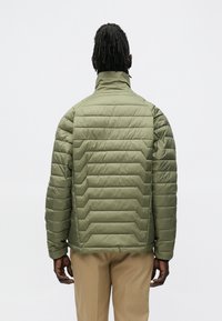 Muž s dredmi v prešívanej olivovozelenej bunde a béžových nohaviciach, stojaci chrbtom k jednoduchému pozadiu.