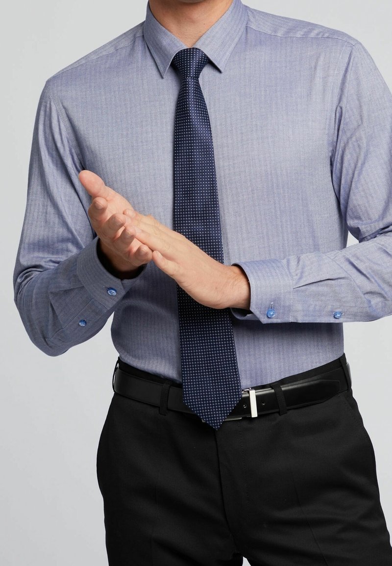 Homme portant une chemise bleue, une cravate sombre à motifs et un pantalon noir, applaudissant devant un fond gris.