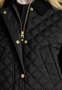 Jachetă neagră matlasată cu accesorii aurii, având un fermoar frontal, guler din corduroy și un buzunar cu petic. Material texturat, ușor.