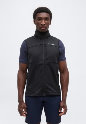 Homme portant un gilet sans manches noir zippé par-dessus un t-shirt bleu marine à manches courtes et un short bleu marine, debout devant un fond uni.