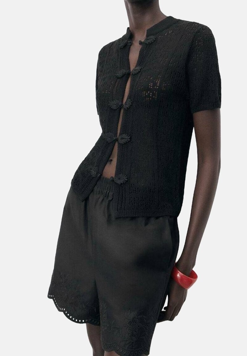 Schwarze gestrickte Kurzarm-Cardigan mit Knopfverschlüssen, kombiniert mit schwarzen Shorts mit gewellten Kanten und floralen Stickereien.