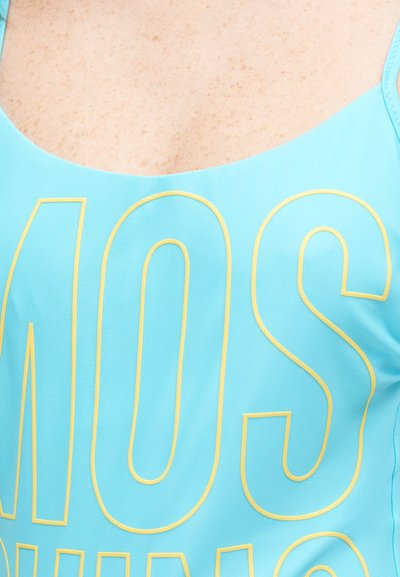 Κοντινό πλάνο ενός ατόμου που φορά ανοιχτό μπλε μπλουζάκι με μεγάλα κίτρινα περιγράμματα γραμμάτων στο στήθος και φαίνεται δέρμα με φακίδες πάνω από το ντεκολτέ.