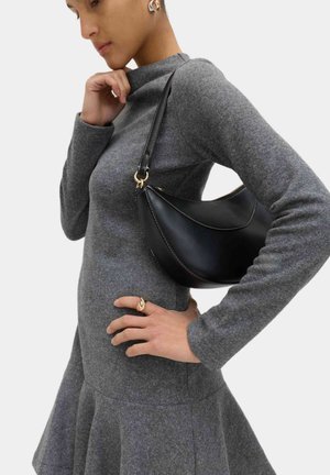 Frau trägt ein langärmliges, graues Strickkleid mit Rüschensaum, hält eine schwarze Leder-Umhängetasche mit goldfarbenen Beschlägen und weißen Nähten.