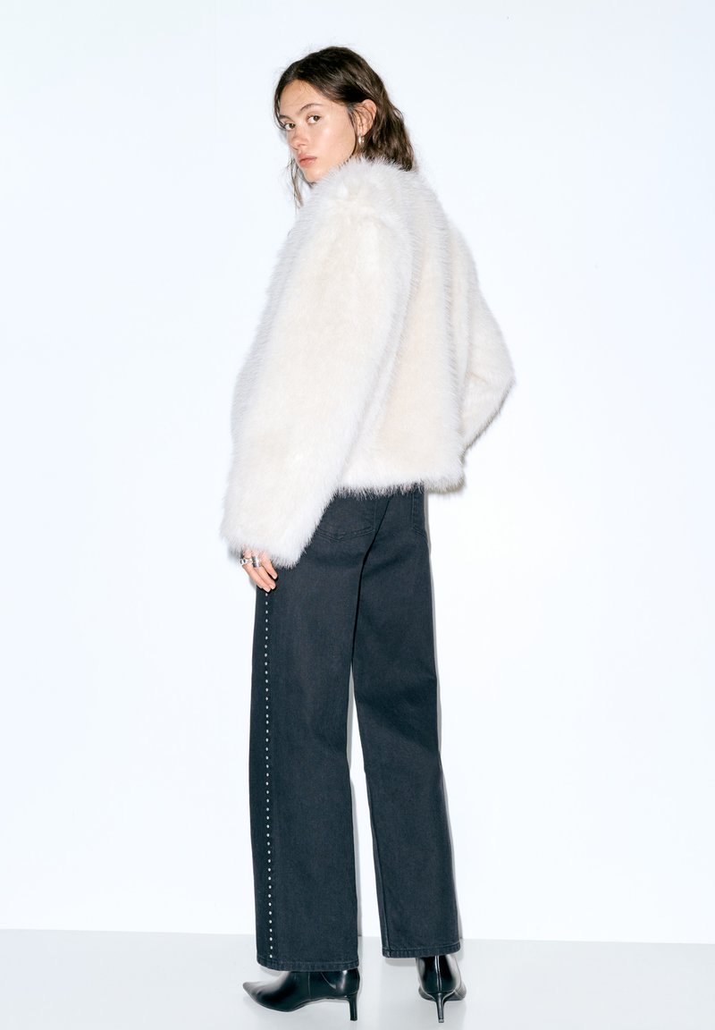 Jachetă albă din blană sintetică, cu decolteu rotund, mâneci lungi și textură pufoasă. Asortată cu pantaloni negri, cu talie înaltă, prevăzuți cu ținte argintii.