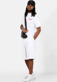 Witte t-shirt met rode graphic, witte shorts, zwarte schoudertas en witte sneakers. Het model draagt een zwarte pet. Eenvoudige, casual outfit.