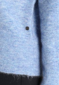 Ljust blå, fluffig tröja med en mjuk textur och en lätt glans. Har en liten mörk knappdetalj på sidan.