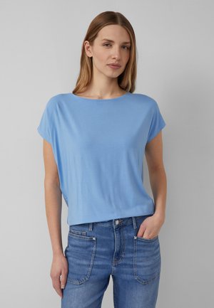 Vrouw met steil lichtbruin haar, een lichtblauw T-shirt met korte mouwen en blauwe jeans, staand met één hand in haar zak tegen een grijze muur.