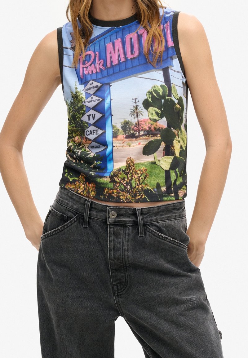 Femme portant un haut court sans manches avec une impression colorée de panneau de motel, cactus, plantes et route, associée à un jean noir taille haute.