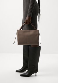 Braune Lederhandtasche mit geflochtenem Griff, rechteckige Form, glatte Textur, kombiniert mit schwarzen knielangen Stilettos und einem figurbetonten Rippkleid.