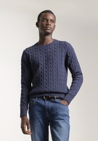 Suéter de punto trenzado azul marino con mangas largas y puños acanalados, combinado con jeans azules. La textura presenta un diseño de patrón de cable prominente.