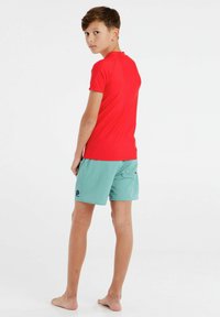 Raudona trumpa rankovėmis marškinėliai, suderinti su akvamarino spalvos trumpikėmis paplūdimiui. Trumpikės turi užpakalinį kišenę ir logotipo dizainą. Modelis stovi basi.