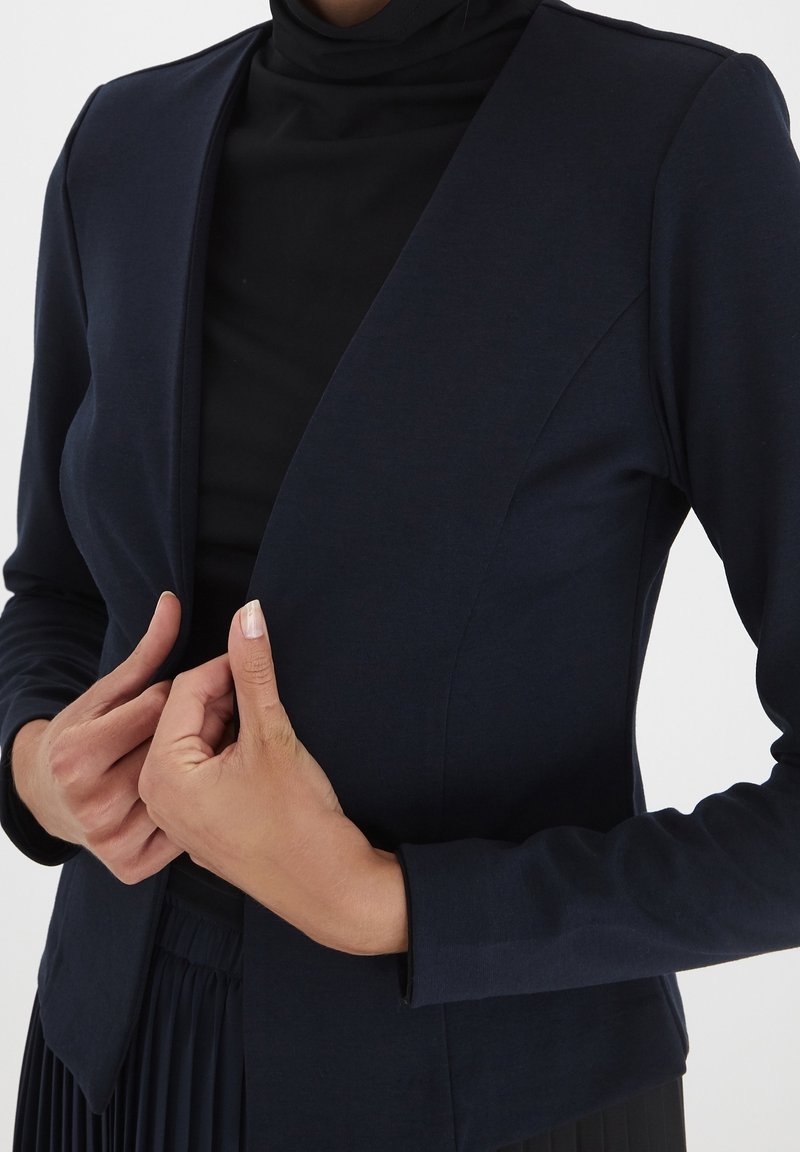 Personne portant un blazer bleu marine foncé sur un haut noir, ajustant le devant du blazer avec les deux mains.