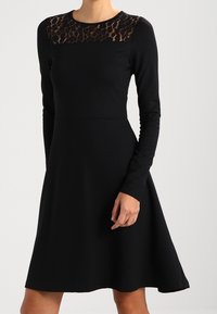 Abito nero a maniche lunghe con corpetto aderente e gonna svasata, caratterizzato da un pannello in pizzo trasparente sul colletto con un sottile motivo leopardato.