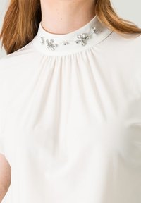 Witte blouse met hoge kraag, versierd met bloemmotieven van heldere kristallen. Zachte stof met delicate plooien rond de halslijn.