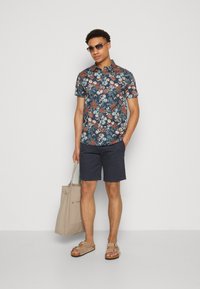 Kortærmet, marineblå skjorte med blomsterprint i rød og blå, parret med marineblå shorts, beige totebag og lyse sandaler.