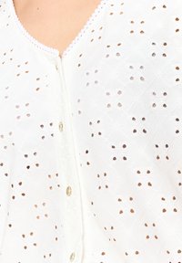 Blusa bianca ricamata con un motivo ad occhio di pernice testurizzato, caratterizzata da un collo scallop, bottoni e piccoli motivi floreali sparsi.