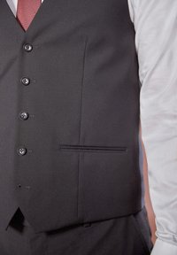 Mustapaita, joka on valmistettu sileästä kankaasta. Siinä on kuusi nappia, yksi rintatasku ja hienovarainen vaakasauma. Paita on puettu valkoisen paidan päälle.