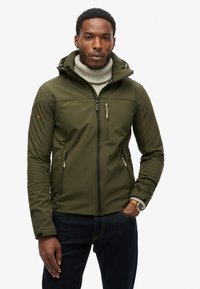 Zielono-oliwkowa kurtka softshell z kapturem, zamykana na zamek błyskawiczny, posiada kieszeń na piersi oraz kieszenie boczne, z teksturowanym wnętrzem i kontrastującymi zamkami.