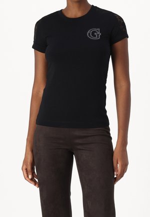 Vrouw draagt een nauwsluitend zwart kortärmelig topje met kant op de schouders en een strass "G" embleem op de linkerborst, gecombineerd met een donkere broek.