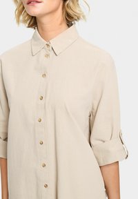 Beige button-up shirt with a soft texture, featuring a collar and rolled sleeves. Brass buttons add detail along the front placket.