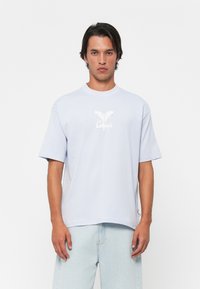 Mann med lyseblå Colcci t-skjorte og lysvaskede jeans, stående mot en enkel hvit bakgrunn.