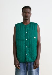 Green quilted vest with snap buttons, featuring two front pockets and a logo label. Sleeveless design, smooth texture, minimal stitching detail.