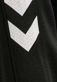 Sort stof med to hvide chevronstriber med et subtilt gentaget "HK"-mønster, syet detalje langs sømmen på højre side.