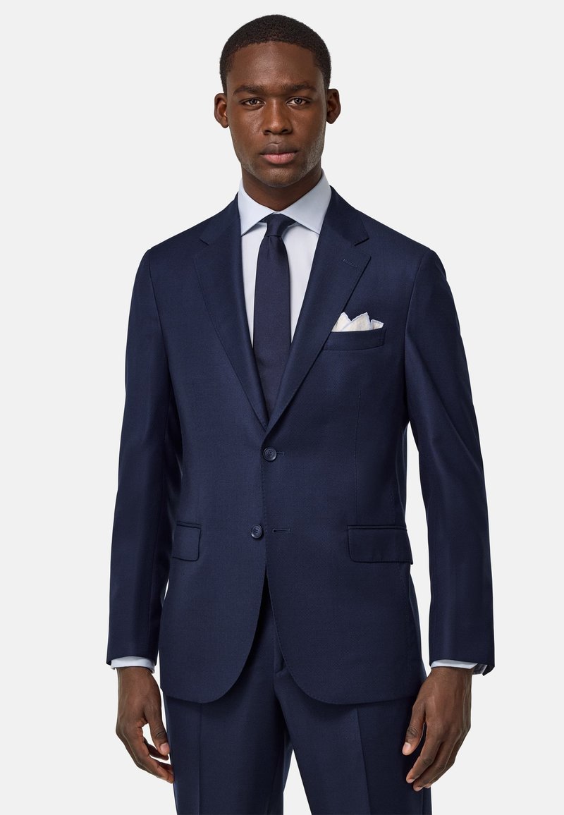 Costume bleu marine avec un revers cranté, deux boutons et des poches avant. Comprend un mouchoir de poche blanc et une cravate sombre assortie. Texture lisse.