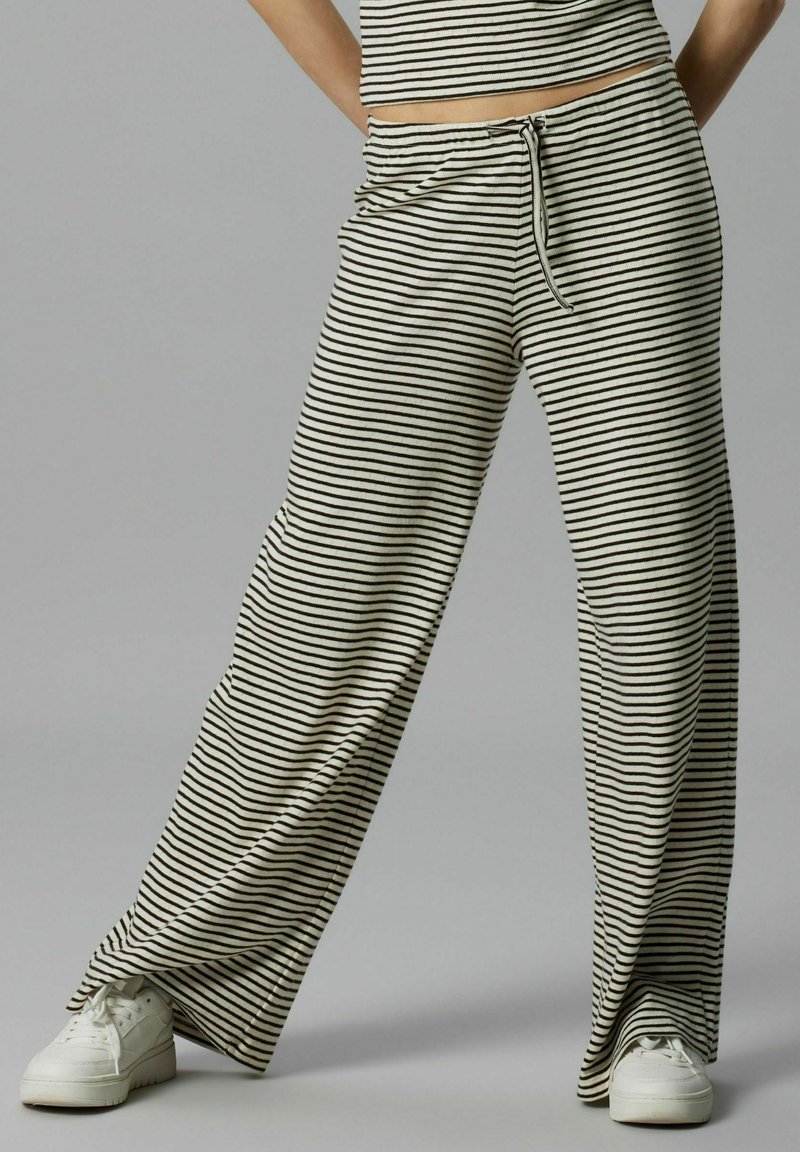 Pantalons larges rayés en tissu beige et noir, avec une taille élastique à cordon de serrage et une coupe décontractée, associés à des baskets blanches.