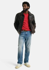 Giacca di pelle marrone con bottoni, camicia rossa, jeans blu sbiaditi e sneakers bianche con dettagli beige. Il modello sta in piedi con le mani in tasca.