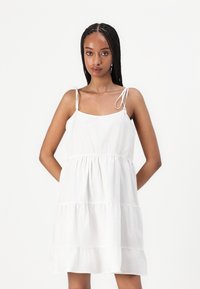 Sleeveless white cotton dress with adjustable tie straps, tiered ruffles at hem, and lightweight, textured fabric.