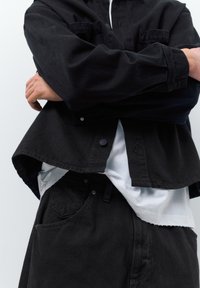 Person iført sort denimjakke og bukser med hvid skjorte under, armene krydset, overkroppen synlig mod ensfarvet baggrund.