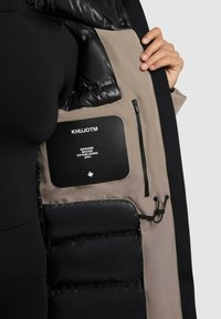 Person holding open a jacket showing a gray interior with black padded sections, zipper pocket, and "KHUJOTM Genuine Brand Established 2001" label.