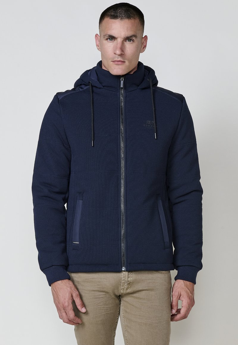 Felpa con zip blu navy realizzata in tessuto testurizzato, con tasche laterali, cappuccio regolabile con cordino e discreta branding sul petto.
