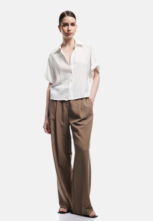 Kobieta z włosami związanymi do tyłu, ubrana w białą koszulę z krótkim rękawem na guziki, luźne brązowe spodnie i czarne sandały, stoi z jedną ręką w kieszeni.