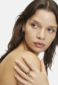 Bague en or rose avec un travail du métal complexe et un design stratifié, portée sur une main contre une épaule. Texture naturelle de la peau et teint légèrement bronzé.