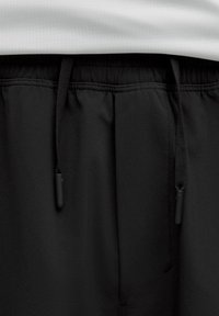 Närbild av svarta elastiska midjebandsbyxor med svarta dragsnören och ett vitt perforerat tyglager ovanför.