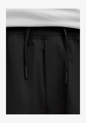 Close-up van zwarte elastische taillebandbroeken met zwarte trekkoorden en een witte geperforeerde stoffenlaag erboven.