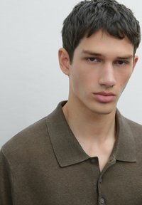 Hnědý pletený polo tričko s límečkem, který má jemnou texturu a dvě knoflíky na výstřihu, na sobě má osoba s krátkými vlasy.