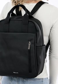 Sac à dos noir en matériau lisse, avec des poignées supérieures et des bretelles réglables. Dispose d'une poche zippée à l'avant et d'un détail de logo.