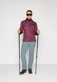 Maroon softshell jacket with zippered pockets, paired with light blue cargo pants and black hiking boots, holding trekking poles.