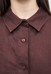 Femme portant une chemise en lin marron avec un col et trois boutons marron visibles, vue du cou à la poitrine.