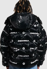 Jachetă puffer neagră cu un finisaj lucios, având logo-uri albe și modele hexagonale. Design cu glugă, textură matlasată și o croială lejeră.
