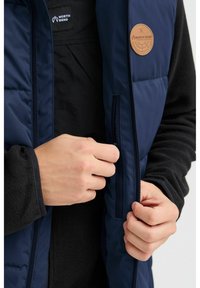 Marinefarbener Puffer-Weste aus glattem Stoff, mit einer Reißverschlusstasche und einem Leder-Logo-Patch. Getragen über einer strukturierten schwarzen Fleece-Schicht.