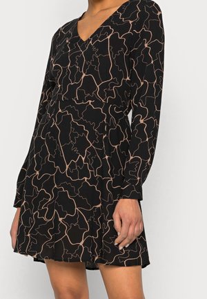 Musta pitkähihainen mekko, jossa beige abstrakti jatkuva viivakuosi, V-pääntie ja levenevä hame, päällä seisovan henkilön.