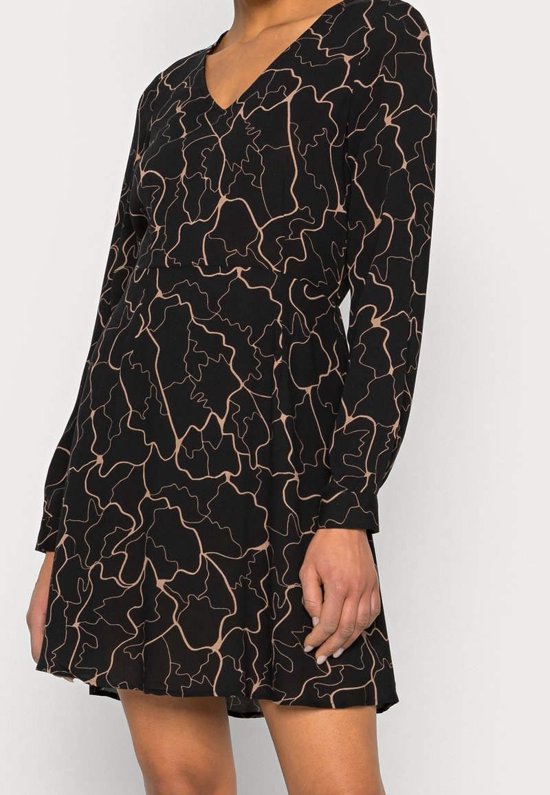 Robe noire à manches longues avec motif abstrait beige en ligne continue, col en V et jupe évasée, portée par une personne debout.