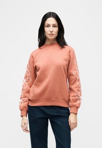 Γυναίκα με φούξια-σαλμον χρώματος φούτερ με λευκά λουλουδάτα σχέδια στα μανίκια και σκούρο μπλε παντελόνι, στέκεται μπροστά σε απλό φόντο.