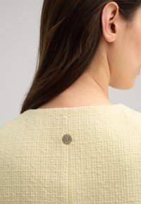 Tessuto giallo texturizzato con un motivo sottile, caratterizzato da un bottone accentato rotondo in argento sul retro del collo.