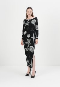 Schwarzes Strickkleid mit langen Ärmeln, das ein weißes Muster aus Muscheln und Seesternen aufweist, seitliche Schlitze und einen Bootsausschnitt hat. Kombiniert mit schwarzen High Heels.