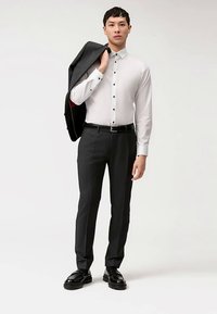 Mannenoutfit met een wit overhemd met zwarte knopen, donker gesneden broek en een zwart colbert over de schouder gedrapeerd.